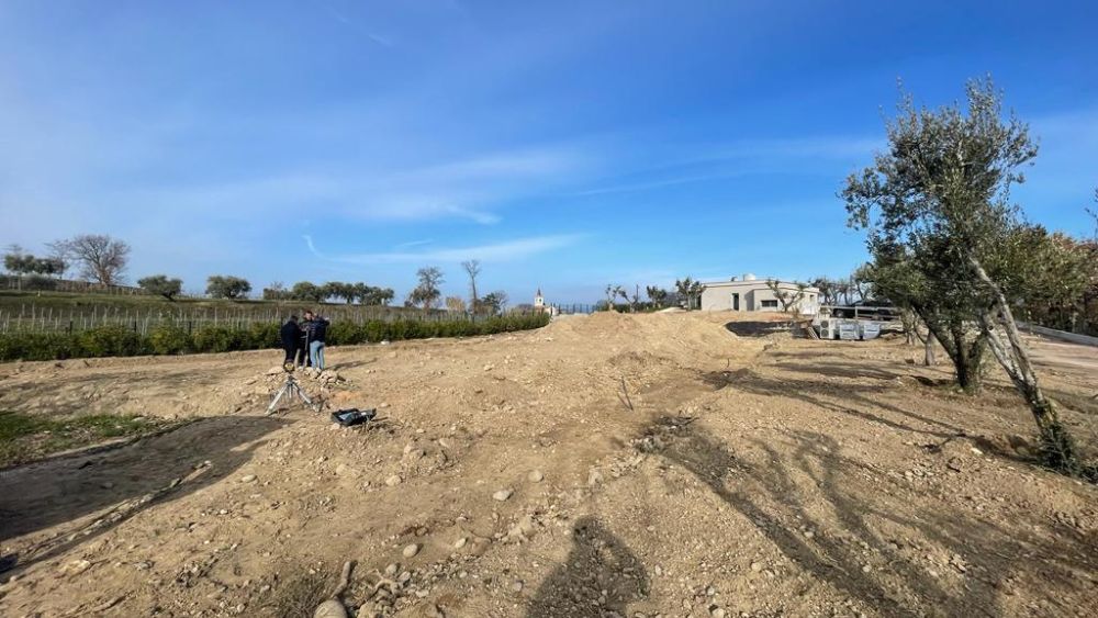 Descrizione: "Questa immagine cattura la trasformazione armoniosa di un terreno agricolo in un moderno impianto sportivo. Al centro, un campo da calcio con erba sintetica verde brillante si distingue nel paesaggio rurale. Le porte regolamentari e le linee di gioco ben definite sul campo sono circondate da elementi agricoli, inclusi campi coltivati e alberi fruttiferi. Una fattoria ristrutturata serve ora come spogliatoio, simbolo di una riconversione rispettosa dell'ambiente. La comunità locale è rappresentata in varie attività, sottolineando il coinvolgimento attivo nella gestione dell'impianto. Dettagli sostenibili come sistemi di raccolta dell'acqua piovana e pannelli solari aggiungono un tocco eco-compatibile. L'immagine riflette un equilibrio perfetto tra sport, agricoltura e sostenibilità, evidenziando l'importanza di impianti sportivi integrati nel tessuto agricolo e comunitario.