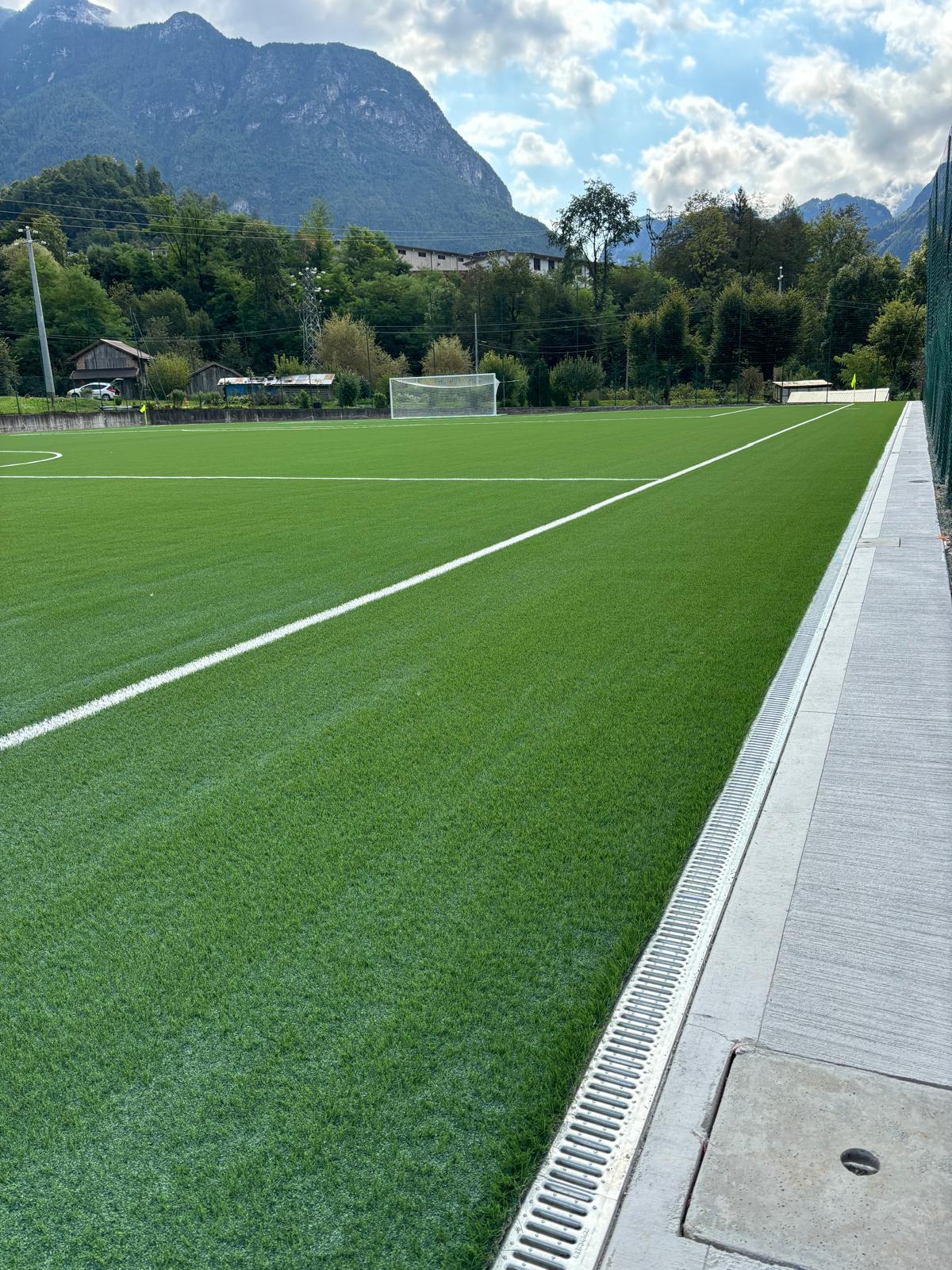 Immagine del nuovo campo da calcio in erba artificiale ad Agordo, caratterizzato da una superficie di gioco uniforme e un sottofondo a drenaggio verticale che garantisce la perfetta gestione dell'acqua piovana e la prevenzione di pozzanghere. Questa soluzione è ideale per il rifacimento campi sportivi, assicurando prestazioni ottimali in qualsiasi condizione atmosferica. Il campo si integra armoniosamente nel paesaggio circostante, contribuendo a creare un impianto sportivo all'avanguardia che risponde alle esigenze della comunità locale.