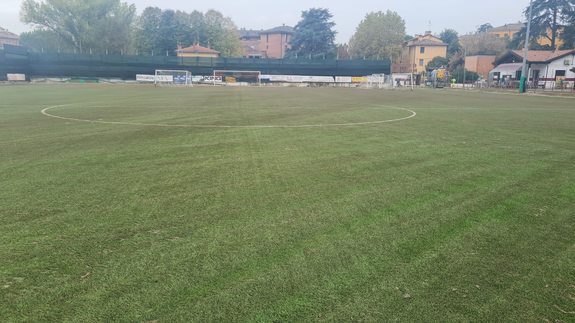 Le immagini mostrano le fasi della manutenzione straordinaria di campi sintetici alluvionati nelle aree colpite dall'alluvione a Bologna. Nella prima immagine, un trattore specializzato è utilizzato per rimuovere e ripristinare la superficie danneggiata del campo sportivo. Nella seconda immagine, si osserva un campo da calcio in fase di recupero, con il manto in erba artificiale in corso di rinnovamento.